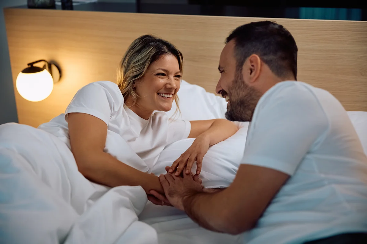 Couple souriant et complice allongé dans un lit aux draps blancs