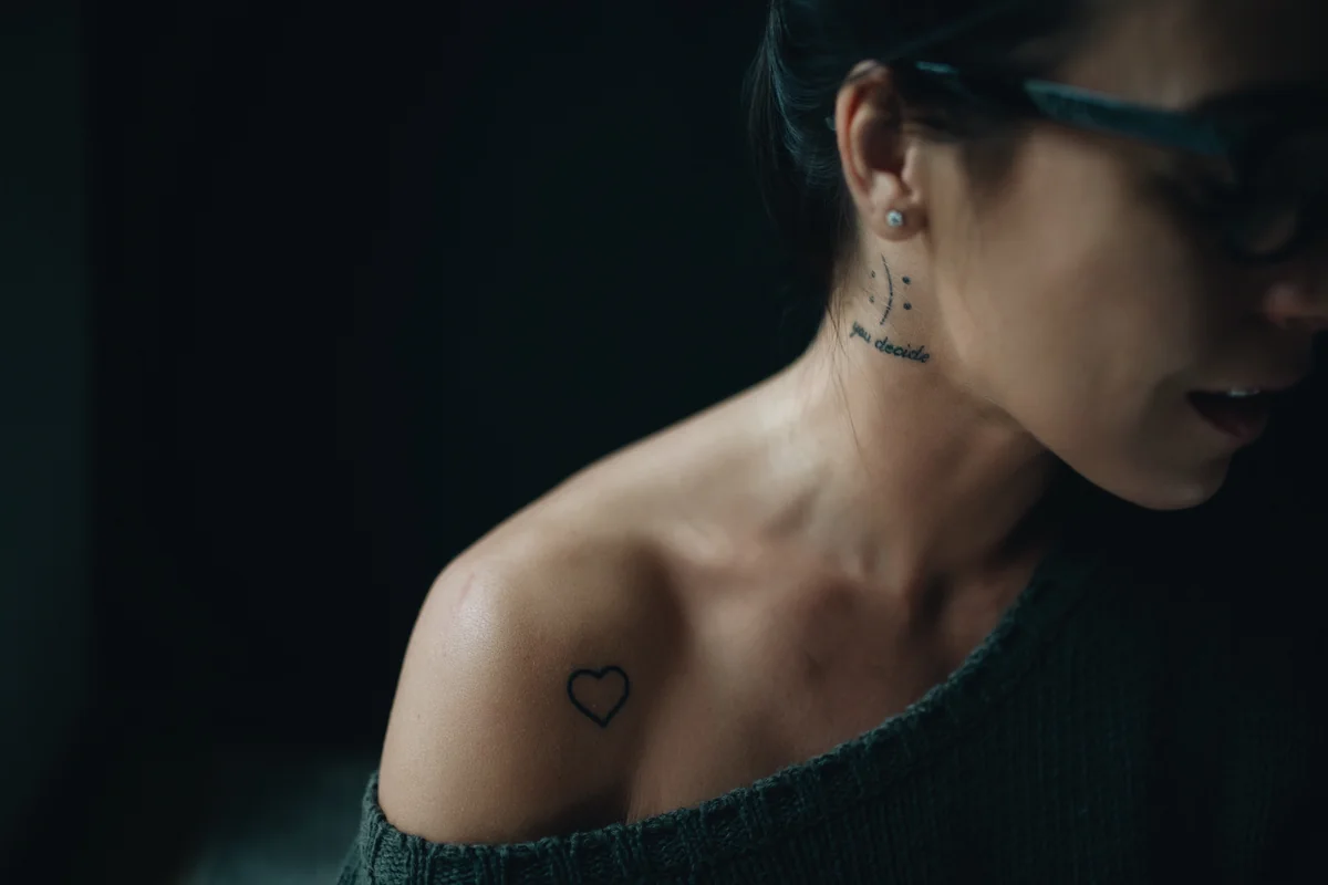 Femme au pull dénudant l'épaule, tatouage cœur sur l'épaule et petit tatouage sur la nuque