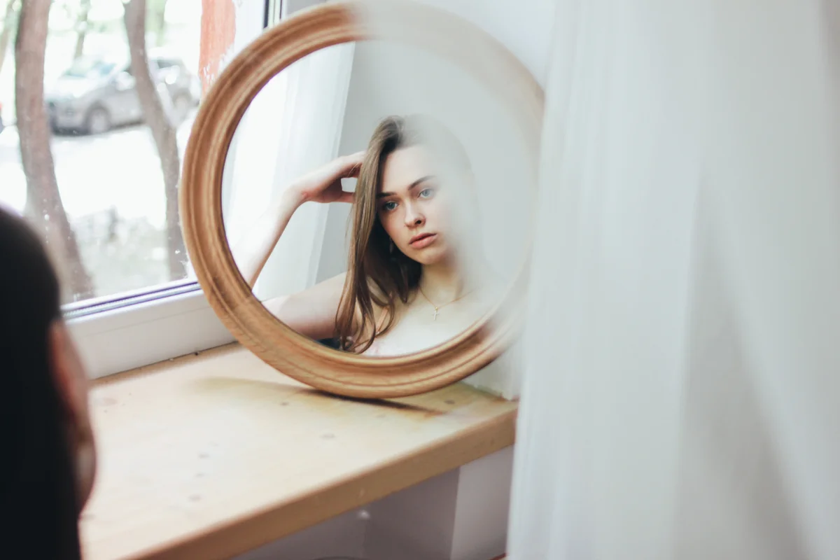 Femme pensive regardant son reflet dans un miroir rond posé près d'une fenêtre