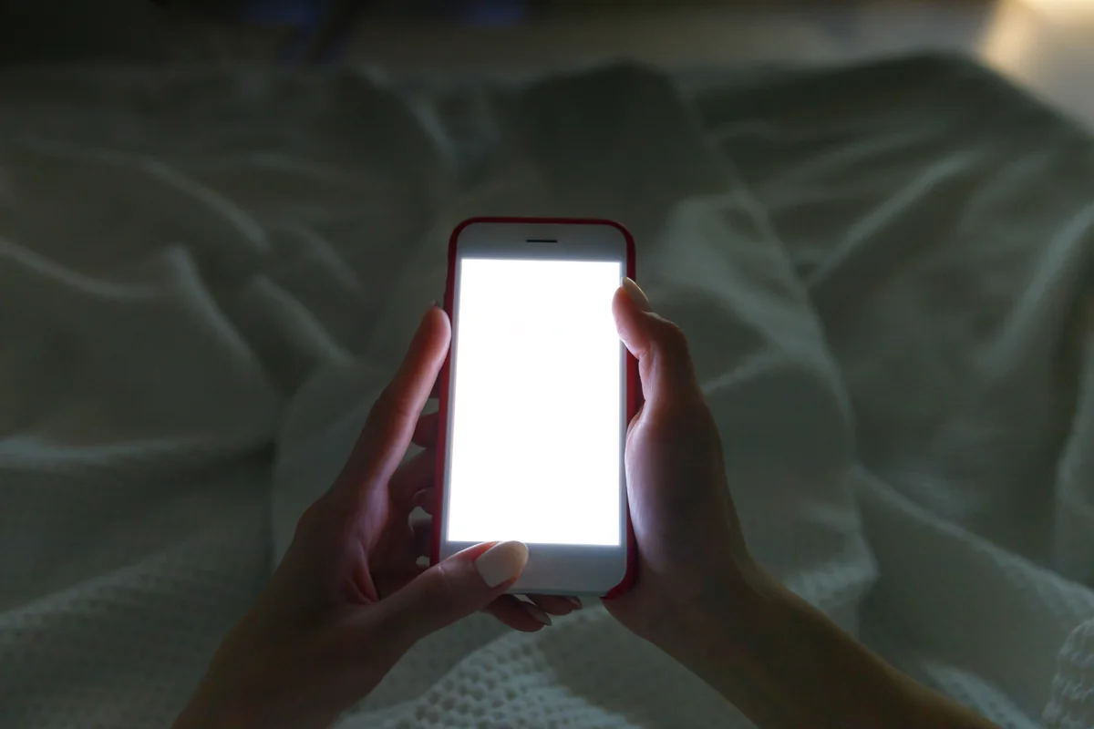 Mains tenant un smartphone allumé au-dessus des draps, la nuit