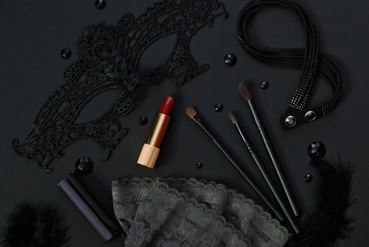 Masque en dentelle noire, rouge à lèvres et pinceaux de maquillage sur fond noir